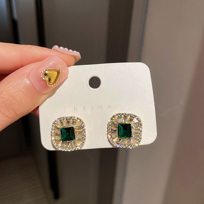 Square-cut emerald earrings with diamond halo settings displayed on white retail card showing luxury jewelry presentation