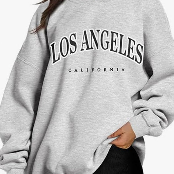 A model wearing an oversized light gray sweatshirt with 'LOS ANGELES CALIFORNIA' text graphic, paired with black leggings on white background