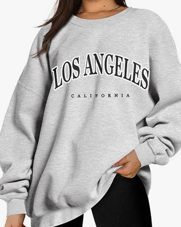 A model wearing an oversized light gray sweatshirt with 'LOS ANGELES CALIFORNIA' text graphic, paired with black leggings on white background