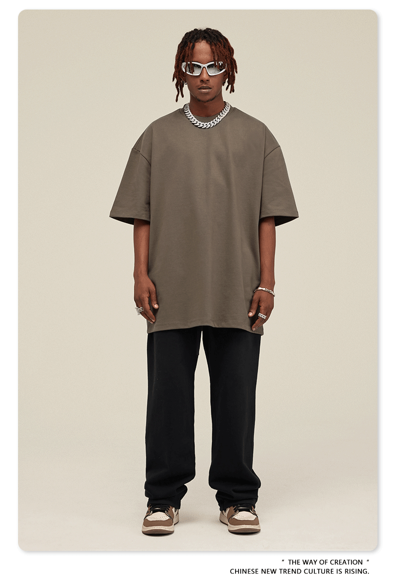Man with short dreadlocks wearing taupe oversized t-shirt, black sweatpants, white sneakers, silver chain necklace and white-framed sunglasses