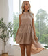 A woman with platinum blonde hair is wearing a taupe halter neck dress and standing by a window with greenery