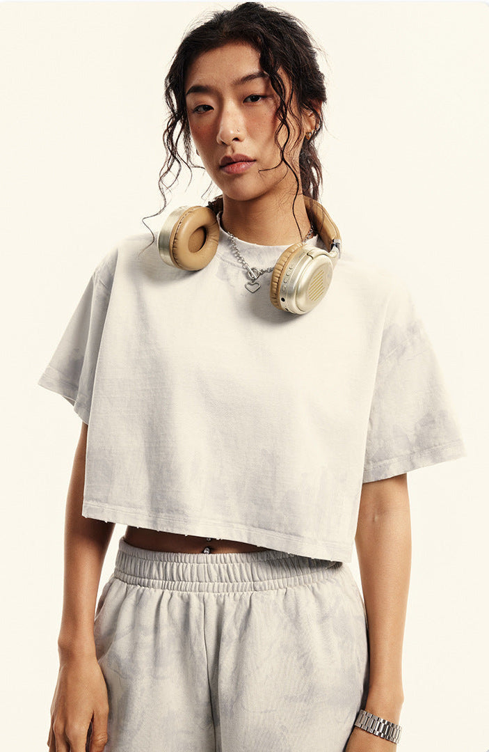 A woman with curly hair wearing gold headphones poses in cream colored crop top and matching shorts against white backdrop
