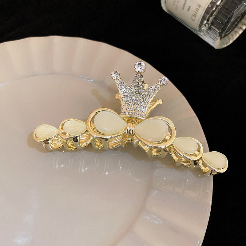 A gold-plated hair clip adorned with a crown-shaped rhinestone embellishment and teardrop-shaped opalescent stones, placed on a white plate.