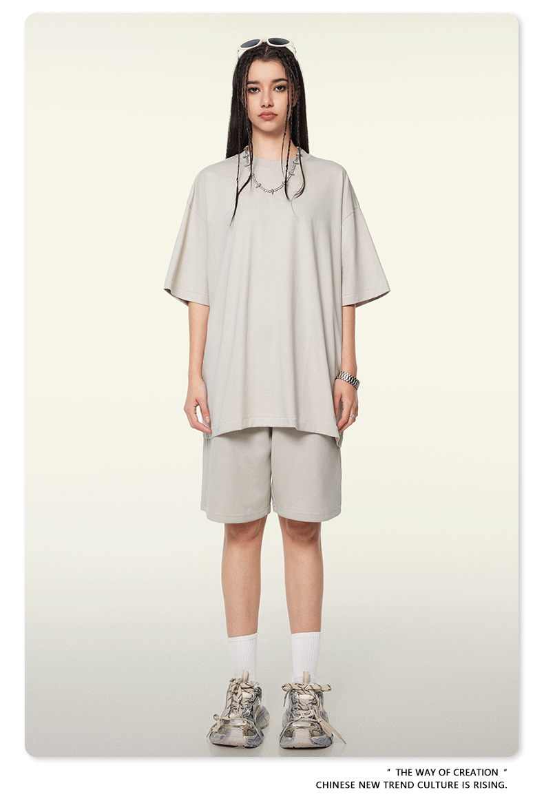 A woman wearing a light beige oversized t-shirt and shorts set, styled with sunglasses on head, silver necklace, and chunky white sneakers against neutral background