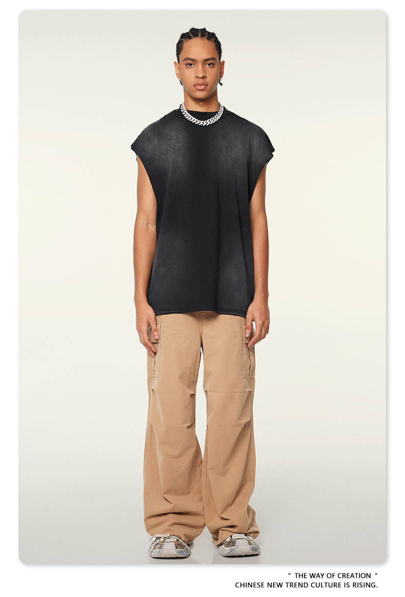 A man wearing a black sleeveless muscle tee and tan cargo pants stands straight against a white background with silver chain necklace