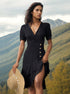 A woman wearing a black wrap midi dress with button details holds a straw hat while posing against a mountainous landscape