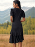 A woman shown from behind wearing a black puff-sleeve dress with layered ruffle hem stands overlooking mountain scenery