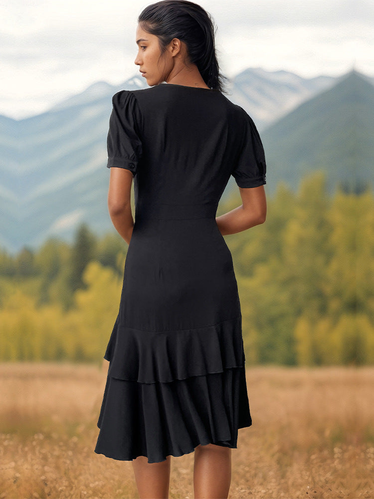 A woman shown from behind wearing a black puff-sleeve dress with layered ruffle hem stands overlooking mountain scenery