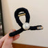 A black looped fabric hair clip featuring a large white pearl and a small engraved metal tag, held in a person’s hand.
