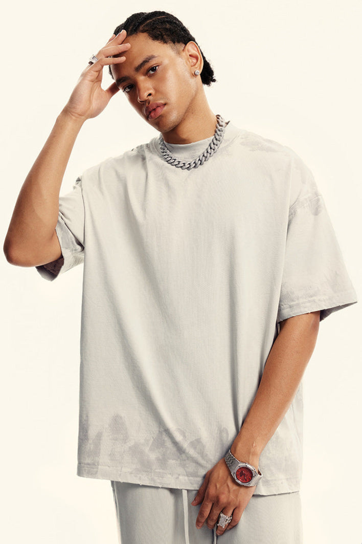 Portrait of model with hand to forehead wearing light beige t-shirt and silver chain. Features minimalist styling with subtle washed fabric effect