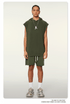 A model wearing olive green sleeveless hoodie with matching shorts, silver pendant necklace and white socks against neutral backdrop