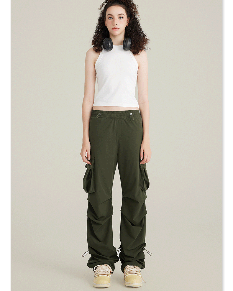 Model in white racerback tank top and olive cargo pants with utility pockets and adjustable drawstring hem. Black headphones rest on shoulders
