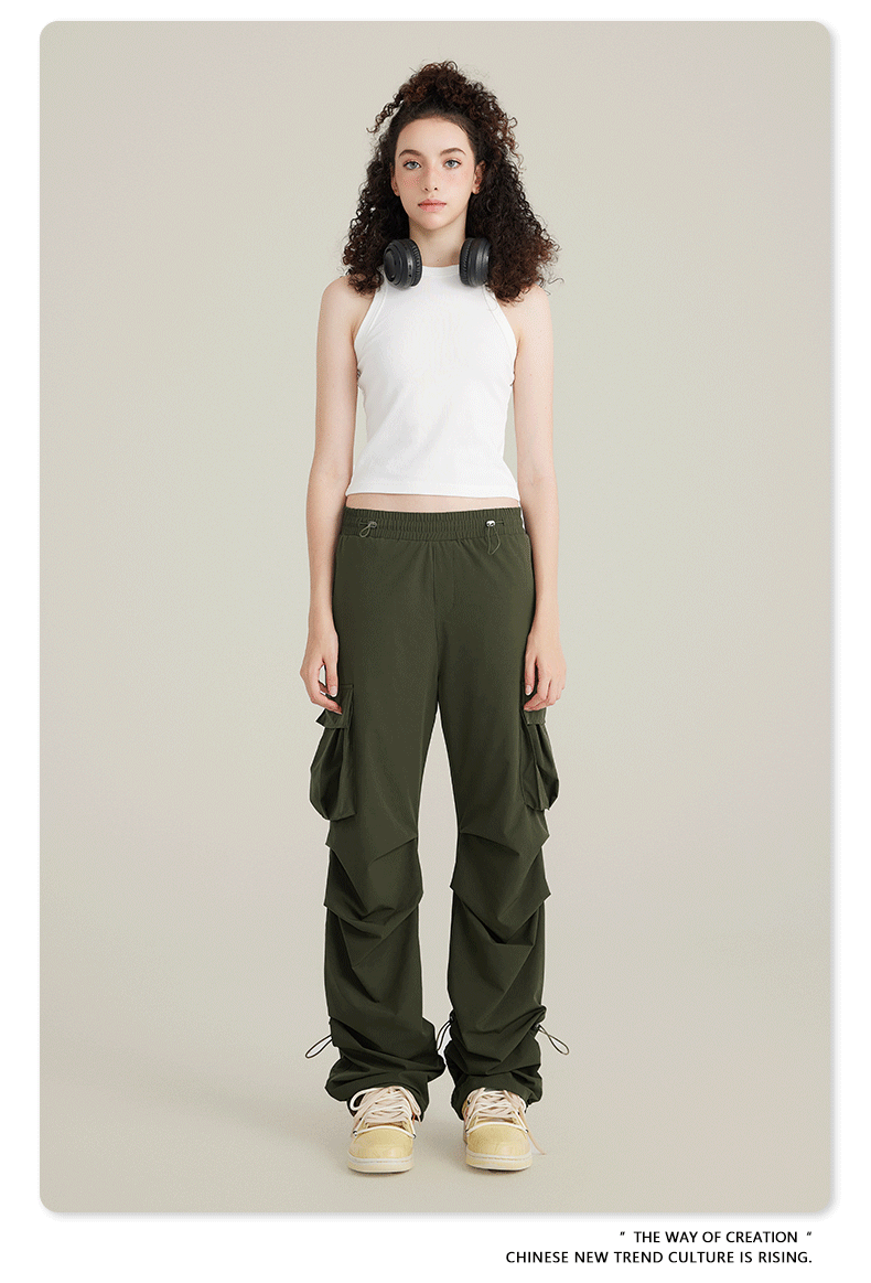 Model in white racerback tank top and olive cargo pants with utility pockets and adjustable drawstring hem. Black headphones rest on shoulders