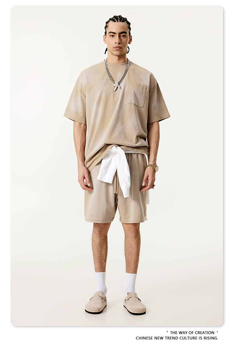 Model in beige t-shirt and shorts with silver necklace, white accent and loafers against cream background