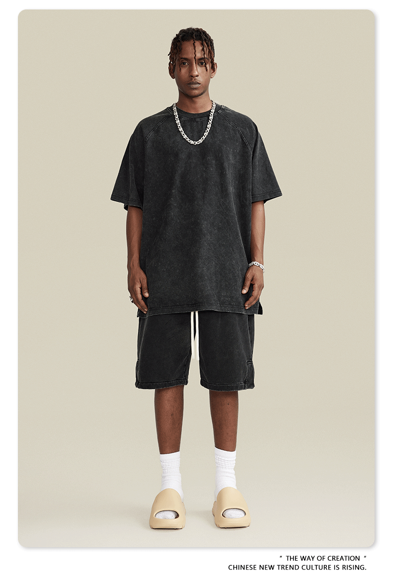 A man wearing an oversized black washed t-shirt, matching knee-length shorts, white socks, beige slide sandals, and a silver chain necklace
