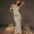 A model is posing with hand behind head wearing an ivory textured gown with corseted bodice and gathered waist against dark studio background