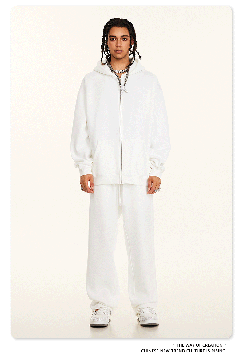 Model wearing light gray oversized zip-up hoodie and matching sweatpants with white rectangular sunglasses and silver chain necklaces against cream background