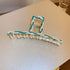 A gold-tone claw clip with turquoise enamel accents and small white pearls, featuring a wire-wrapped design, displayed on a beige surface.