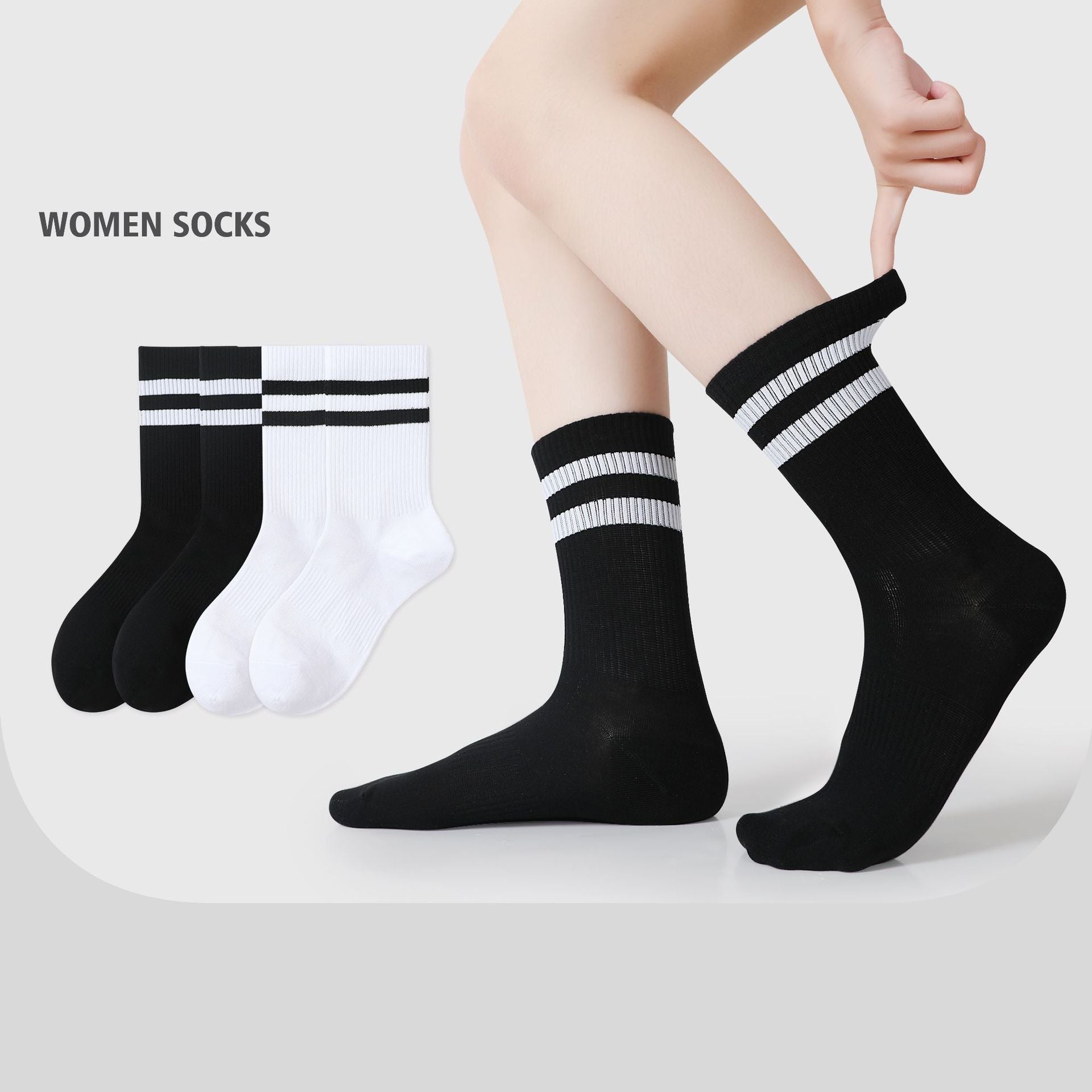 A model is wearing black athletic crew socks with two white stripes, sitting on a white surface while adjusting the sock’s cuff