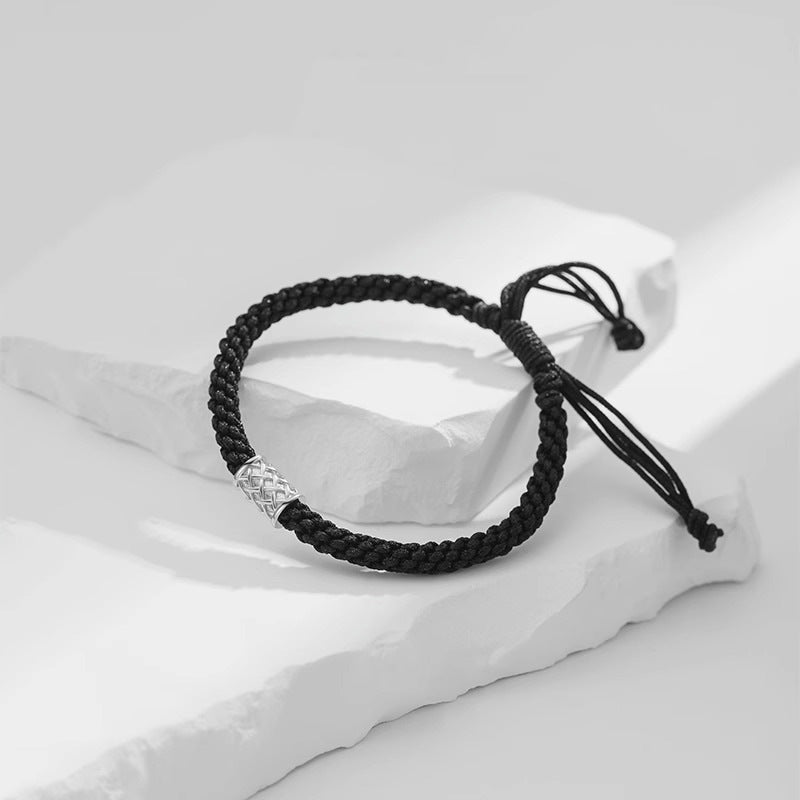 A black braided rope bracelet with a silver cylindrical charm featuring an intricate pattern, displayed on white stone slabs.