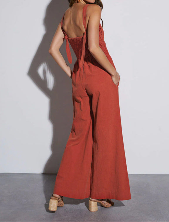 A woman in a rust-colored jumpsuit with an open back and tie straps, standing against a white background.