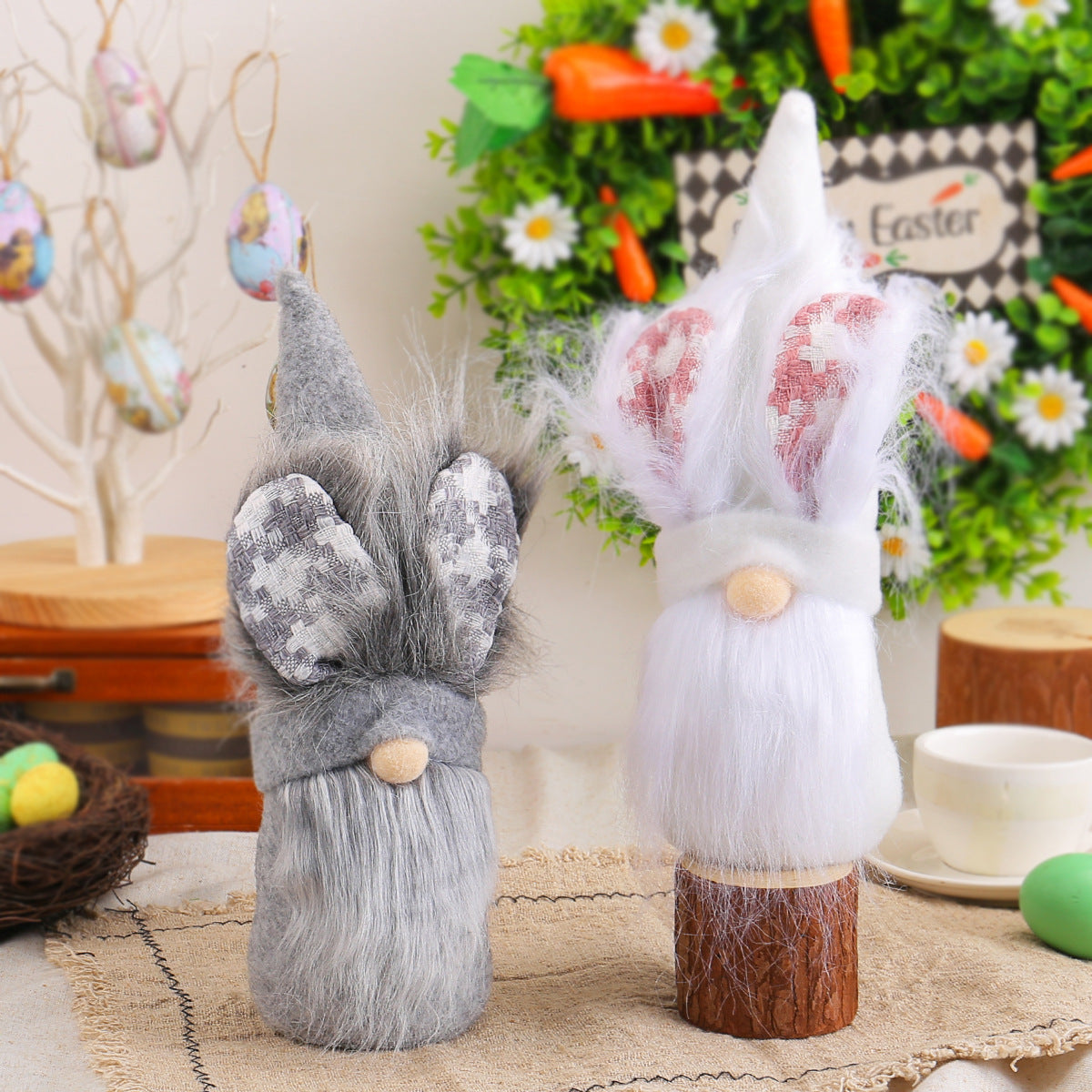 Gray and white Easter gnome plush dolls with bunny ears, posed on a wooden table with a Happy Easter wreath and hanging eggs in the background.