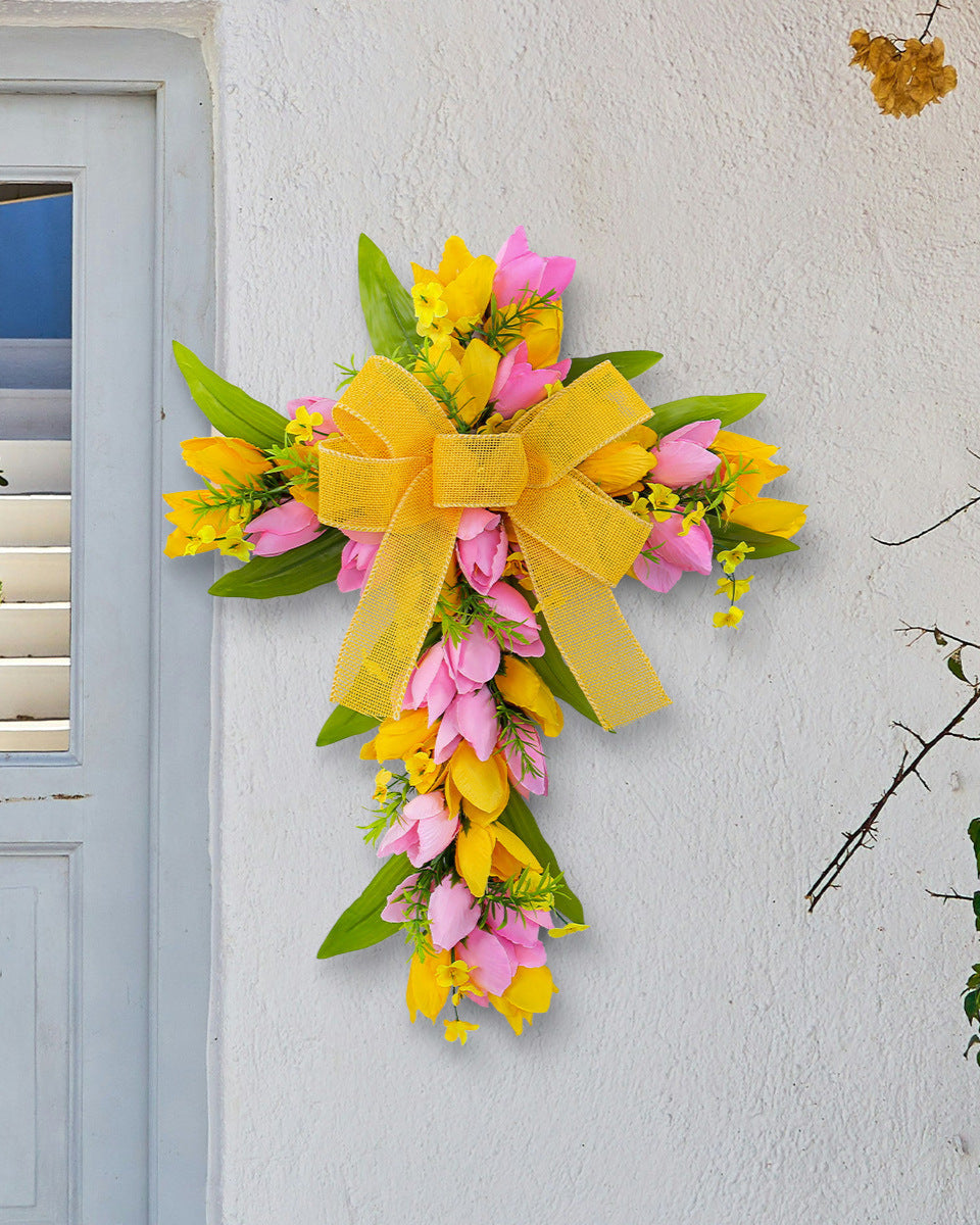 A yellow and pink floral cross wreath with a large yellow bow, mounted on a white wall near a door with natural greenery.