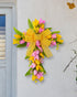 A yellow and pink floral cross wreath with a large yellow bow, mounted on a white wall near a door with natural greenery.