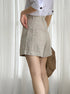 Side view of model wearing beige linen shorts with white button-up shirt and woven bag against sheer curtain backdrop