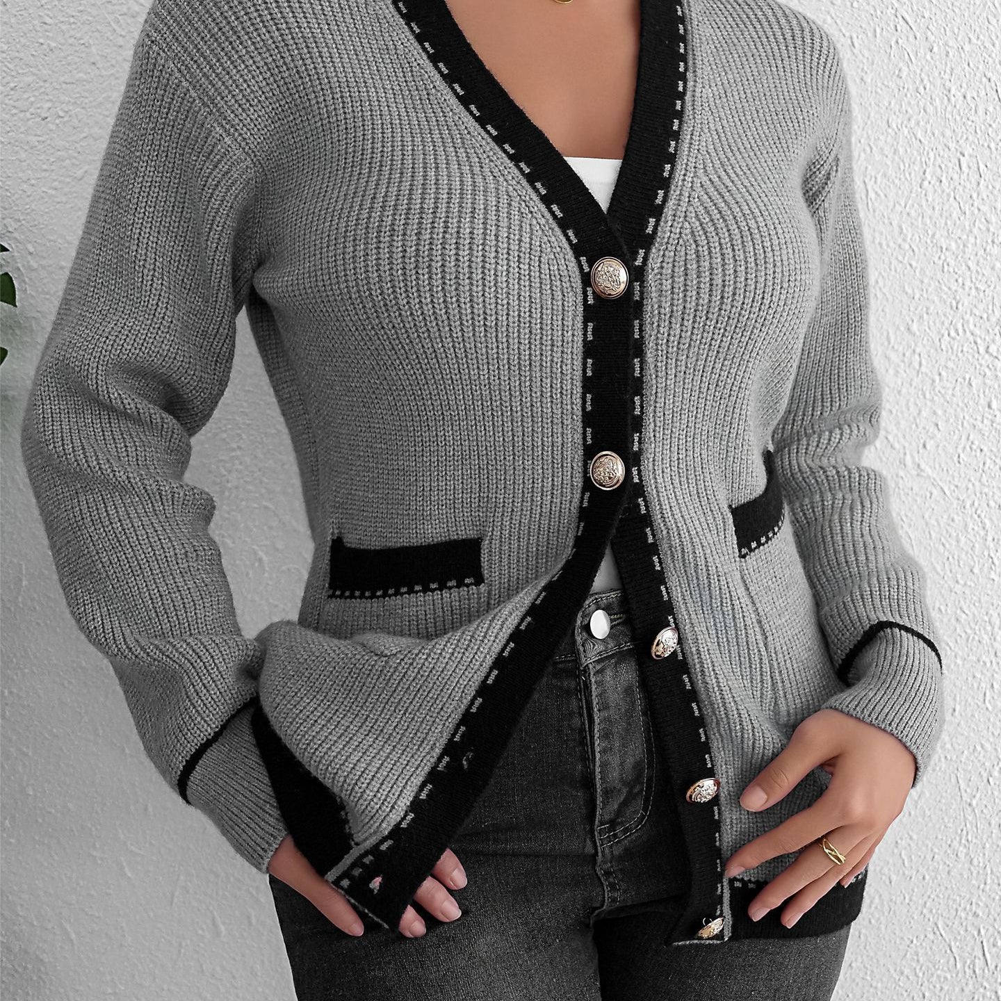Gray knit cardigan with black trim, gold buttons, worn over white top and black jeans, woman standing against white wall