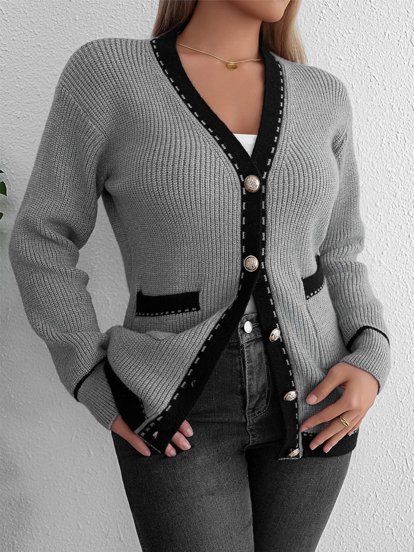 Gray knit cardigan with black trim, gold buttons, worn over white top and black jeans, woman standing against white wall