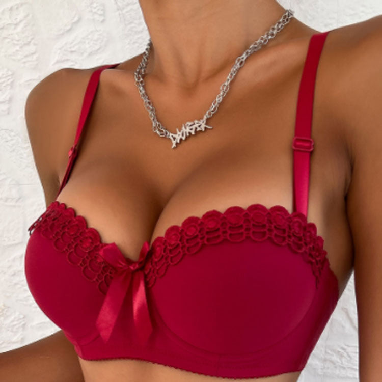 A woman wearing a burgundy lace-trimmed push-up bra with bow detail and silver chain necklace, photographed against white textured background