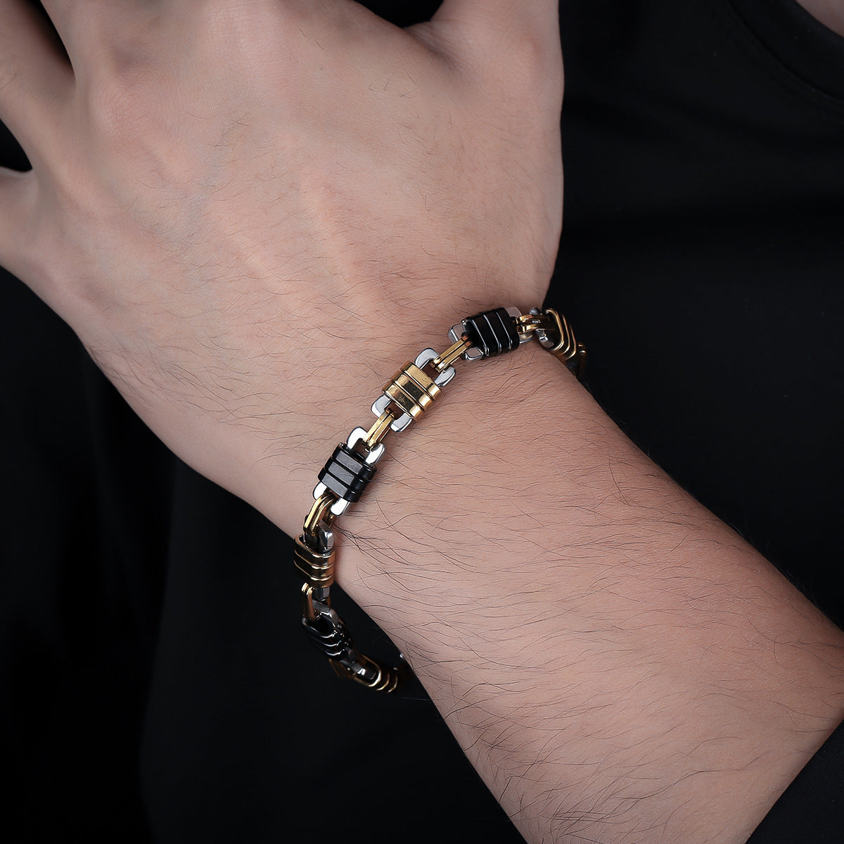 A stylish black and gold stainless steel bracelet with an industrial chain-link pattern displayed on a wrist, paired with a black outfit.
