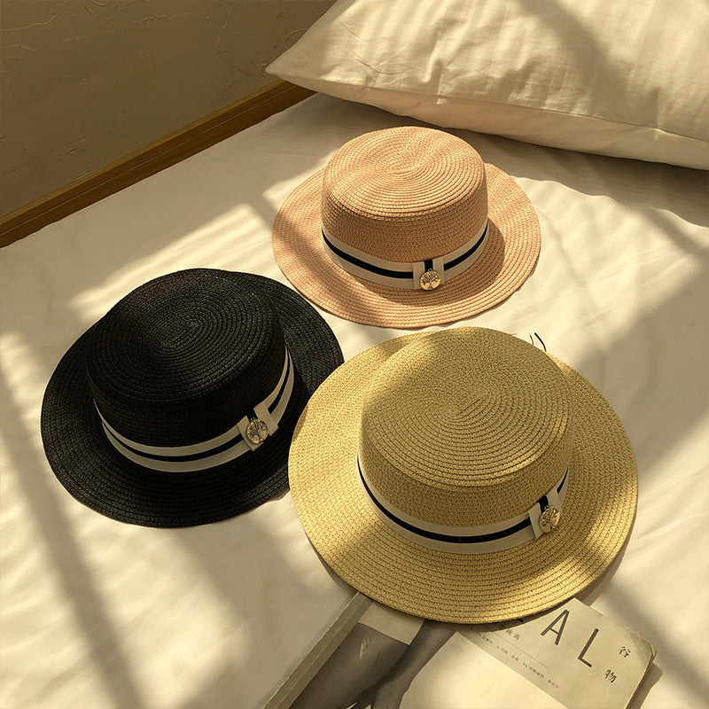 A set of three straw boater hats in black, beige, and pink, featuring a black-and-white ribbon band with a metal tree charm, placed on a white bed.