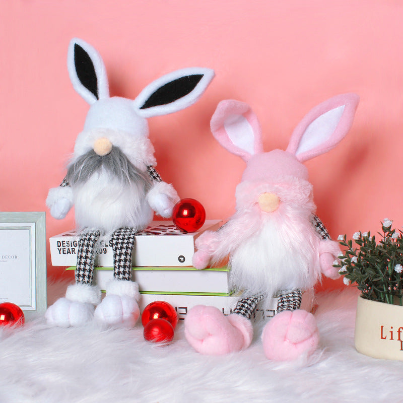 Two Easter bunny gnome dolls, one white and one pink, with long ears and fluffy beards, sitting on books with red ornaments around them.