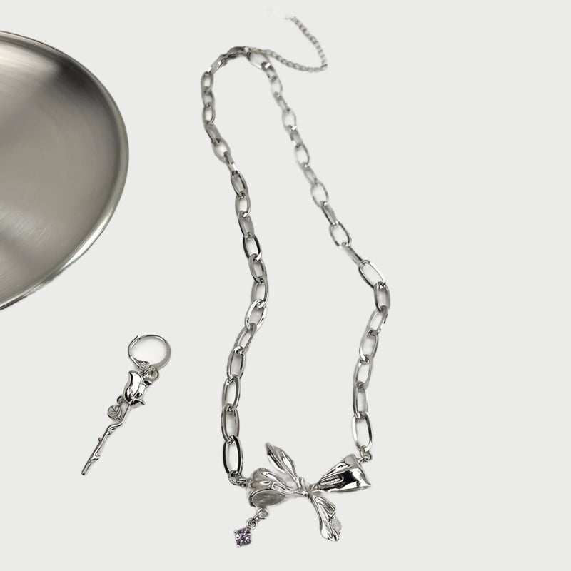 Silver chain necklace with bow charm laid flat beside matching key charm. Features oval links and purple accent stone