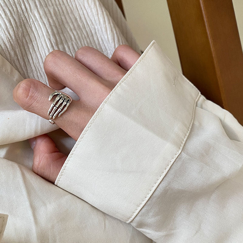 Minimalist shot of a silver skeleton hand ring worn on a person's finger, paired with a neutral outfit