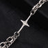 A close-up of a silver chain bracelet featuring a cross pendant with a small gemstone at the center, laid on a black textured surface.