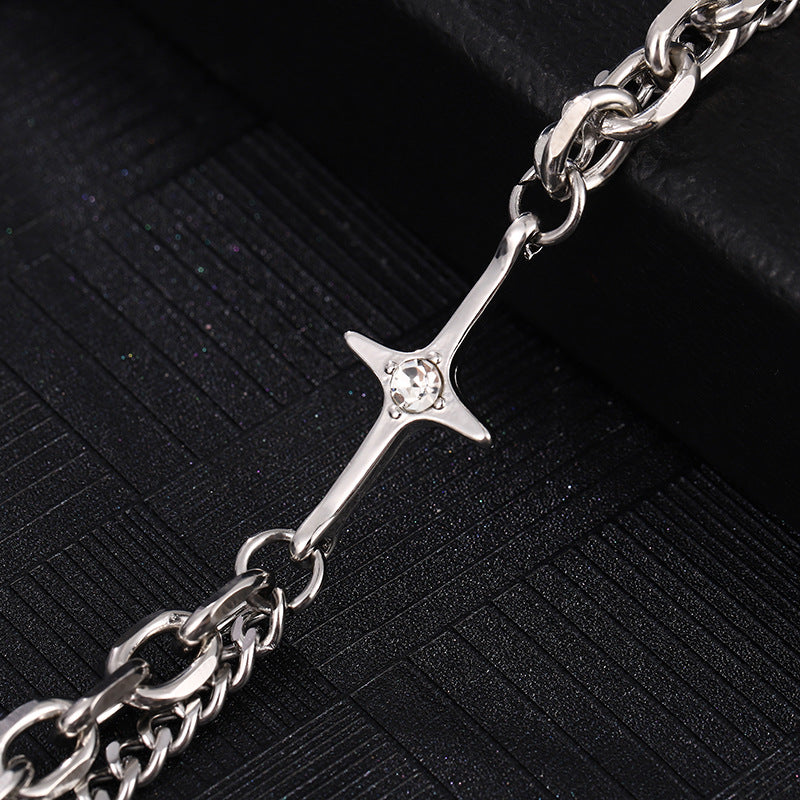 A close-up of a silver chain bracelet featuring a cross pendant with a small gemstone at the center, laid on a black textured surface.