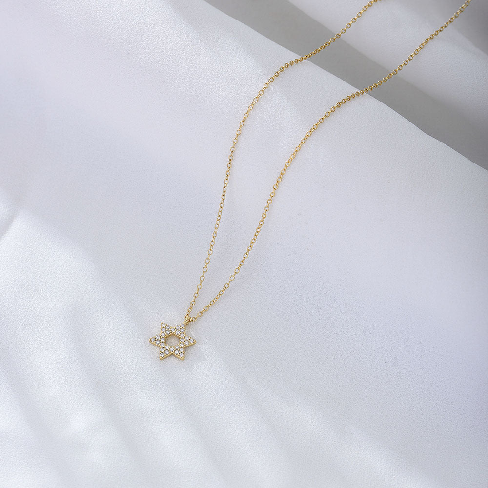 A gold Star of David pendant necklace with diamond-like crystals displayed on white fabric. The elegant six-pointed star hangs on a fine gold chain
