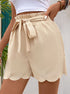 A model wearing beige scalloped hem shorts with elasticated paperbag waist and tie detail against tropical background