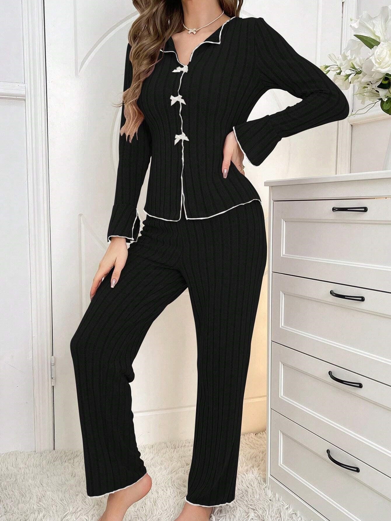 Black ribbed pajama set with white accent trim and bow closures, styled with pearl necklace, shown against white dresser and floral decor