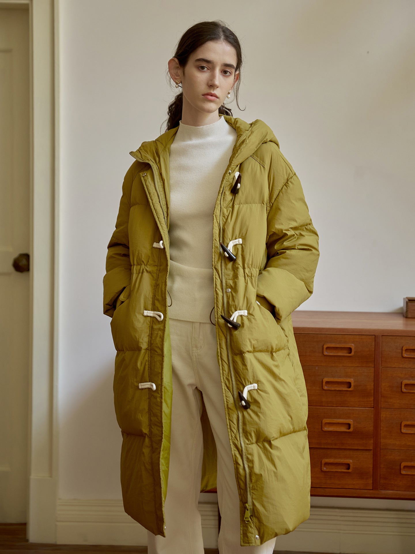 Olive green quilted puffer coat with toggle closures and hood, worn with cream turtleneck and pants