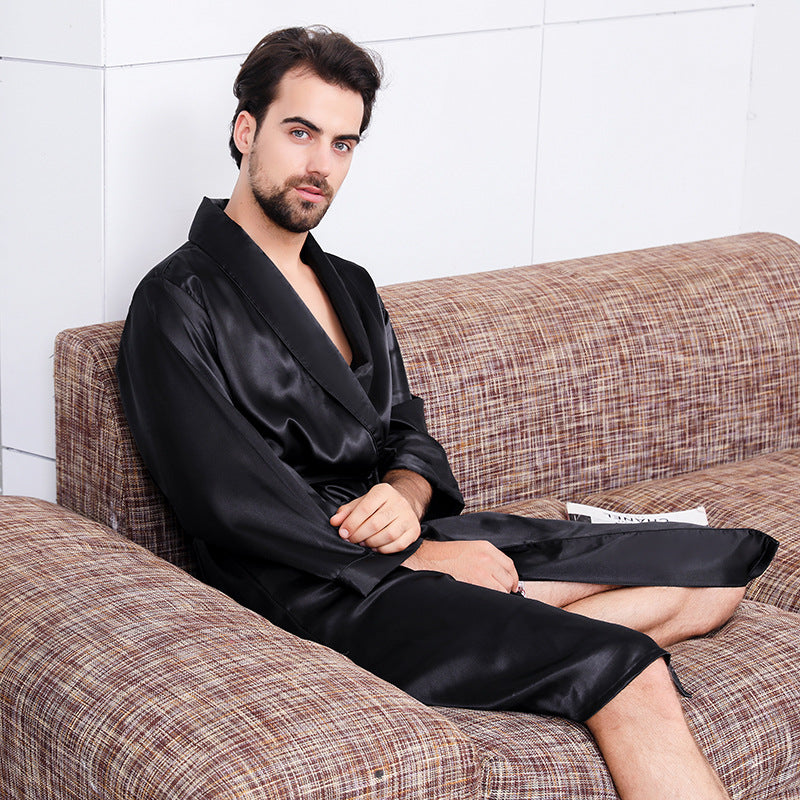 Man seated on a beige tweed sofa wearing an untied black silk robe, looking directly at camera in a minimalist white interior setting
