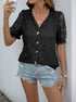 Woman wearing a black lace blouse with intricate floral patterns, styled with denim shorts and accessories, posing against a white wall.