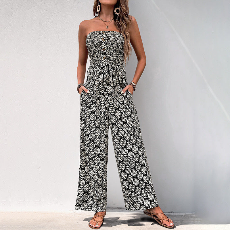 Woman standing against a white wall in a black and white patterned strapless jumpsuit, hands in pockets, wearing layered jewelry.