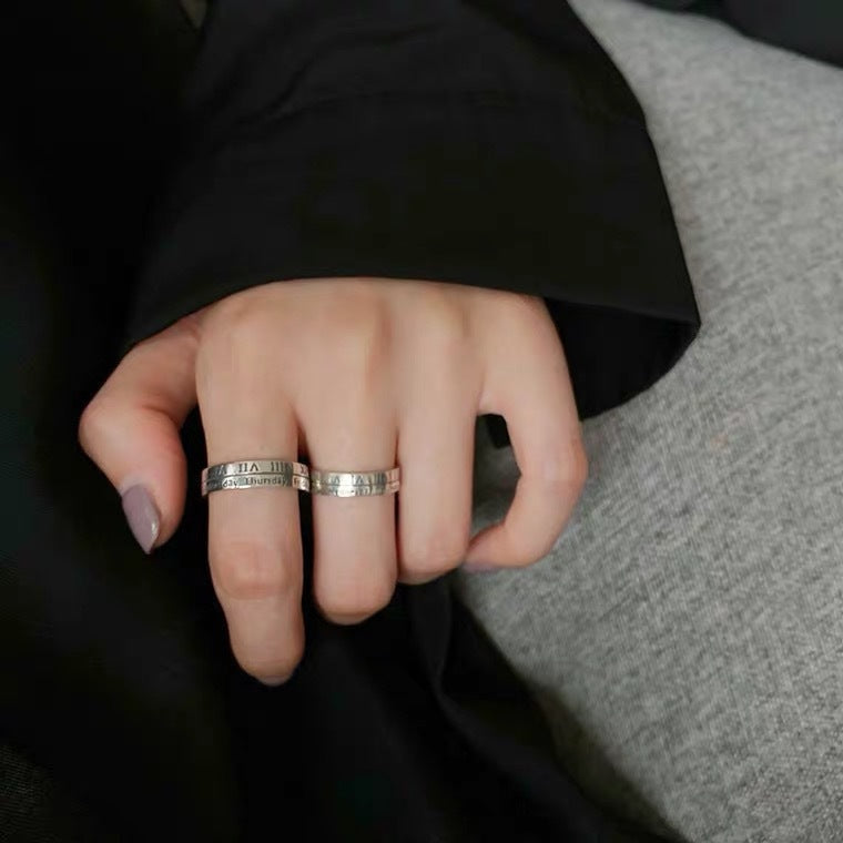 Close-up of hand with mauve nails wearing two matching silver Roman numeral rings with weekday engravings on grey surface