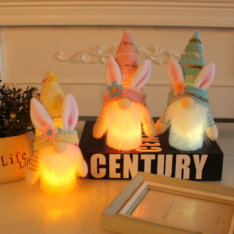  Three LED bunny gnome decorations in pastel colors, glowing warmly on a tabletop with books and decorative accents.