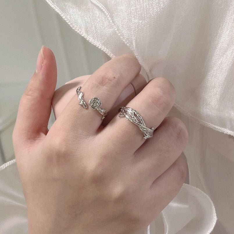 A hand holding white fabric while wearing silver twig-design rings with rose detail, displayed against white satin background
