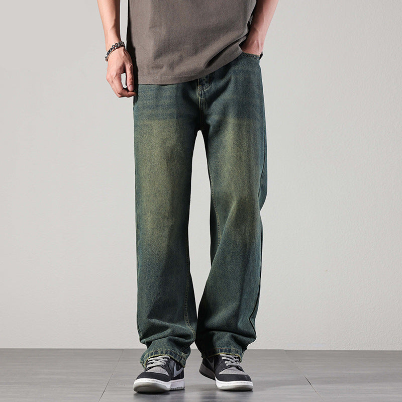A man wearing faded greenish-blue loose-fit jeans, a dark t-shirt, and black sneakers with white soles, standing against a plain background